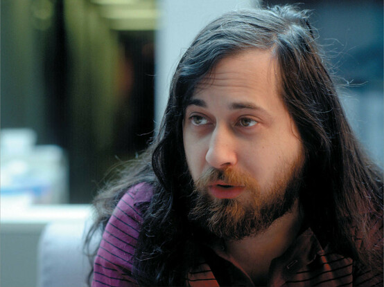 A young Richard Matthew Stallman is shown. He has long hair and a trimmed bread, smooth skin, and is looking away from the camera, perhaps addressing someone. 