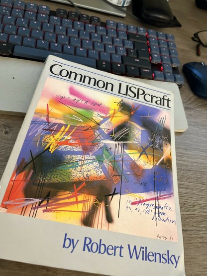 A photo of the book „Common LISPcraft“ by Robert Wilensky laying on a desk. A keyboard, mouse are also visible.