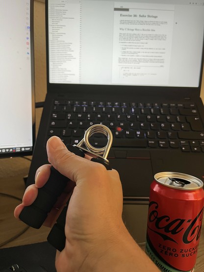 A hand with a Hand Grip Strenghener in front of a ThinkPad X1 laptop, on the screen a page from the book „Learn C The Hard Way“ chapter „Why C Strings Were a Horrible Idea“. Next to it a Coke Zero can.